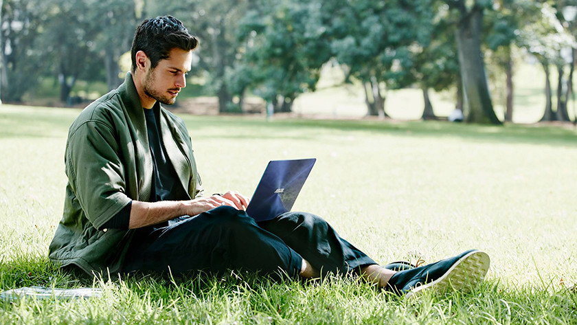 A man sitting in the grass is working on an Asus laptop in the park.